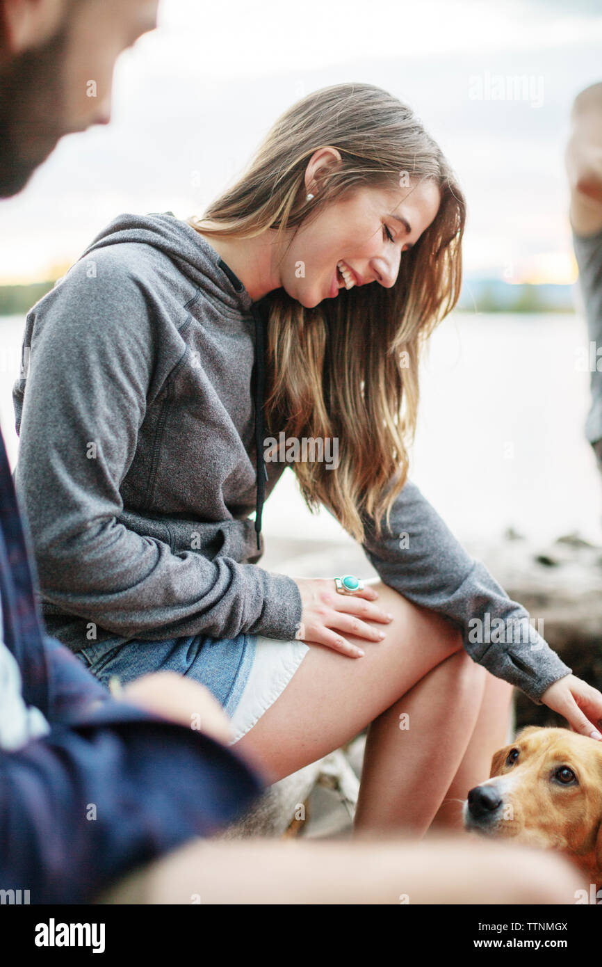 Donna sorridente strofinarsi il cane mentre è seduto da amico contro il fiume Foto Stock