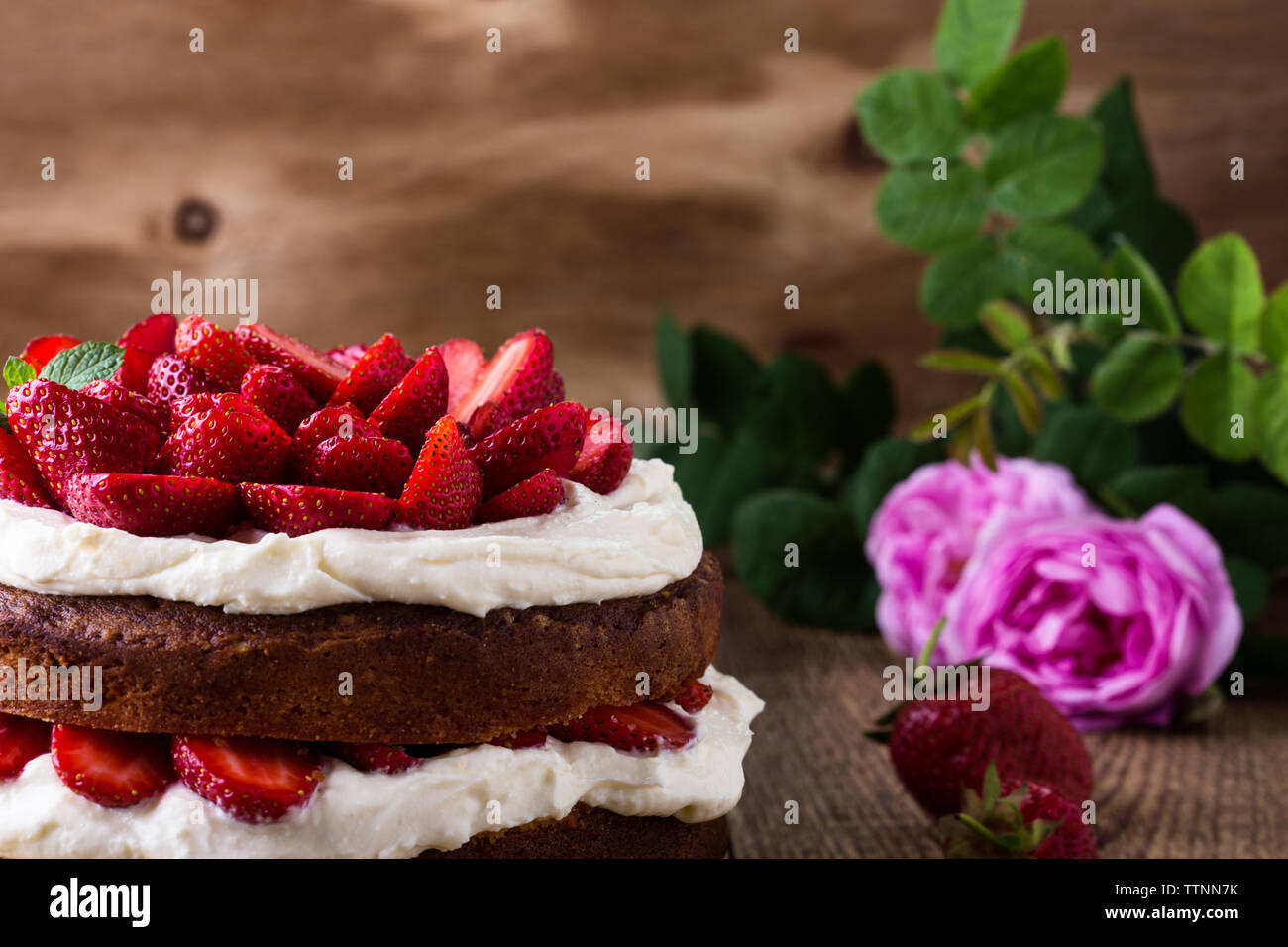 Crema di burro fatti in casa torta con fragole, festosa tabella con le rose rosa e bacche, rustico sfondo di legno Foto Stock