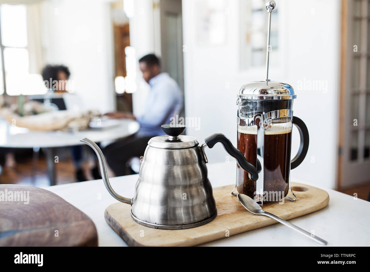 French press e bollitore su tagliere con coppia in background Foto Stock