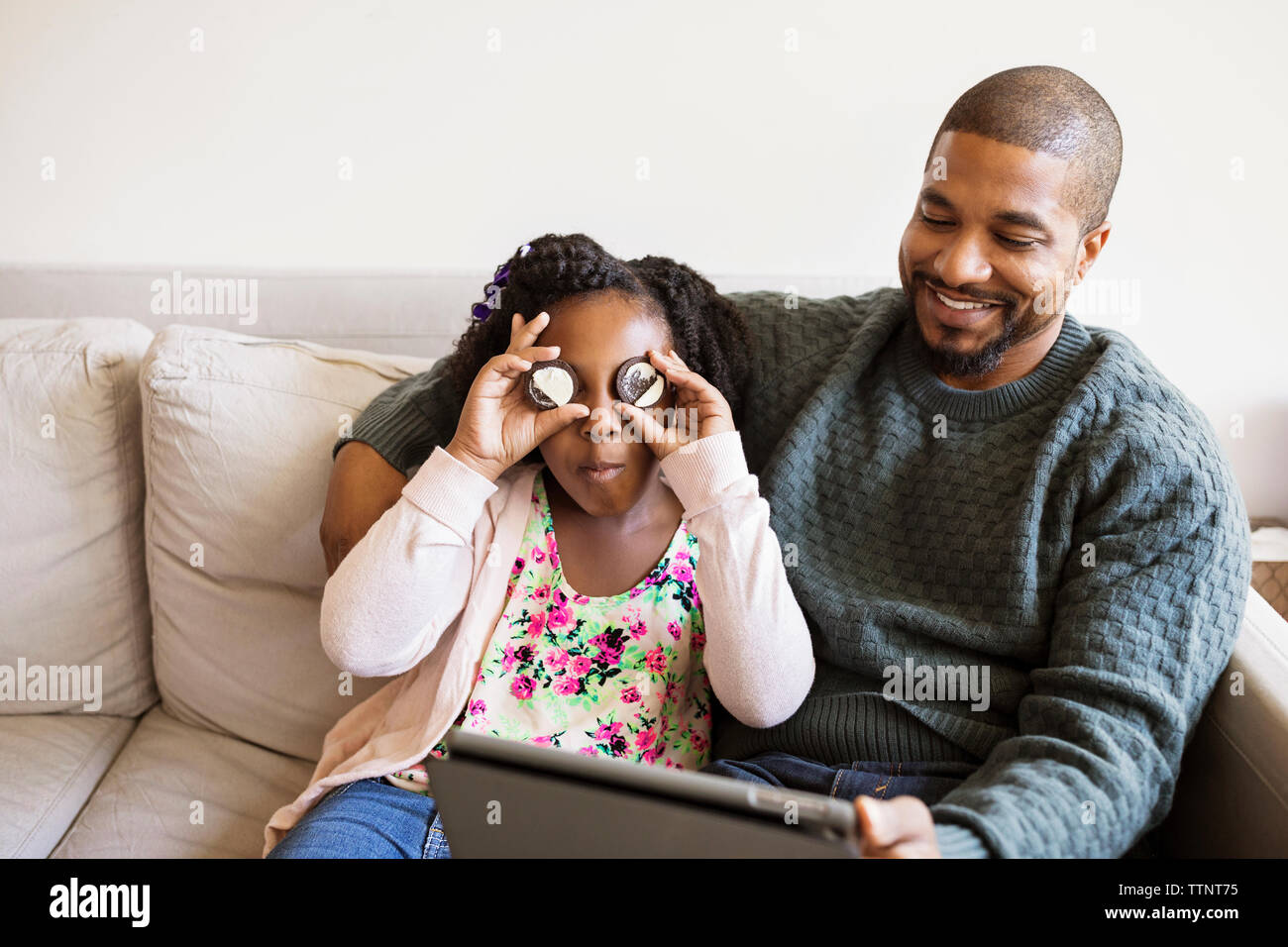 Ragazza che gioca con biscotti mentre è seduto con padre utilizzando computer tablet sul divano Foto Stock