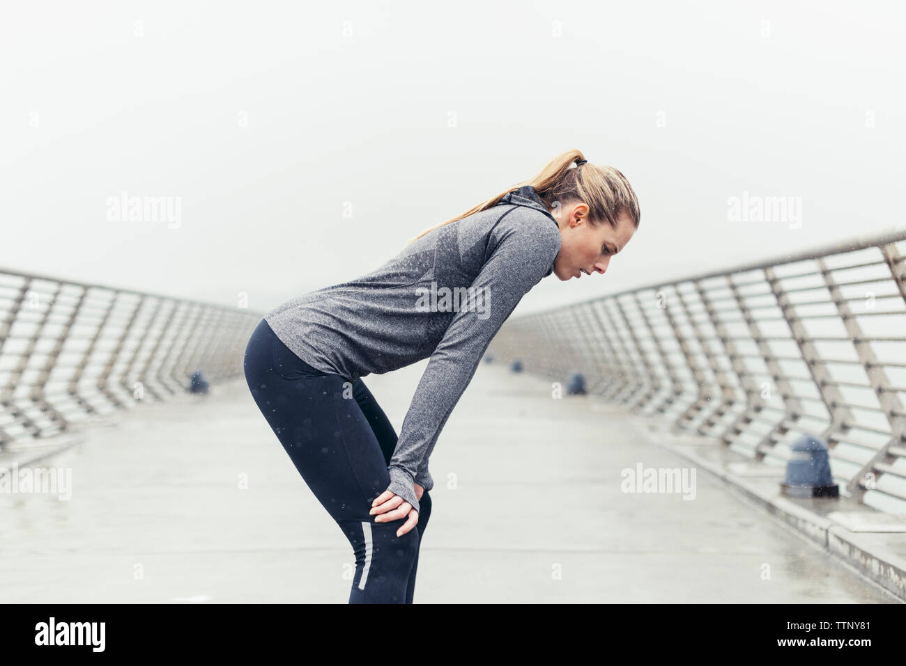 Vista laterale del stanco atleta femminile in piedi con le mani sulle ginocchia sul ponte contro il cielo chiaro Foto Stock