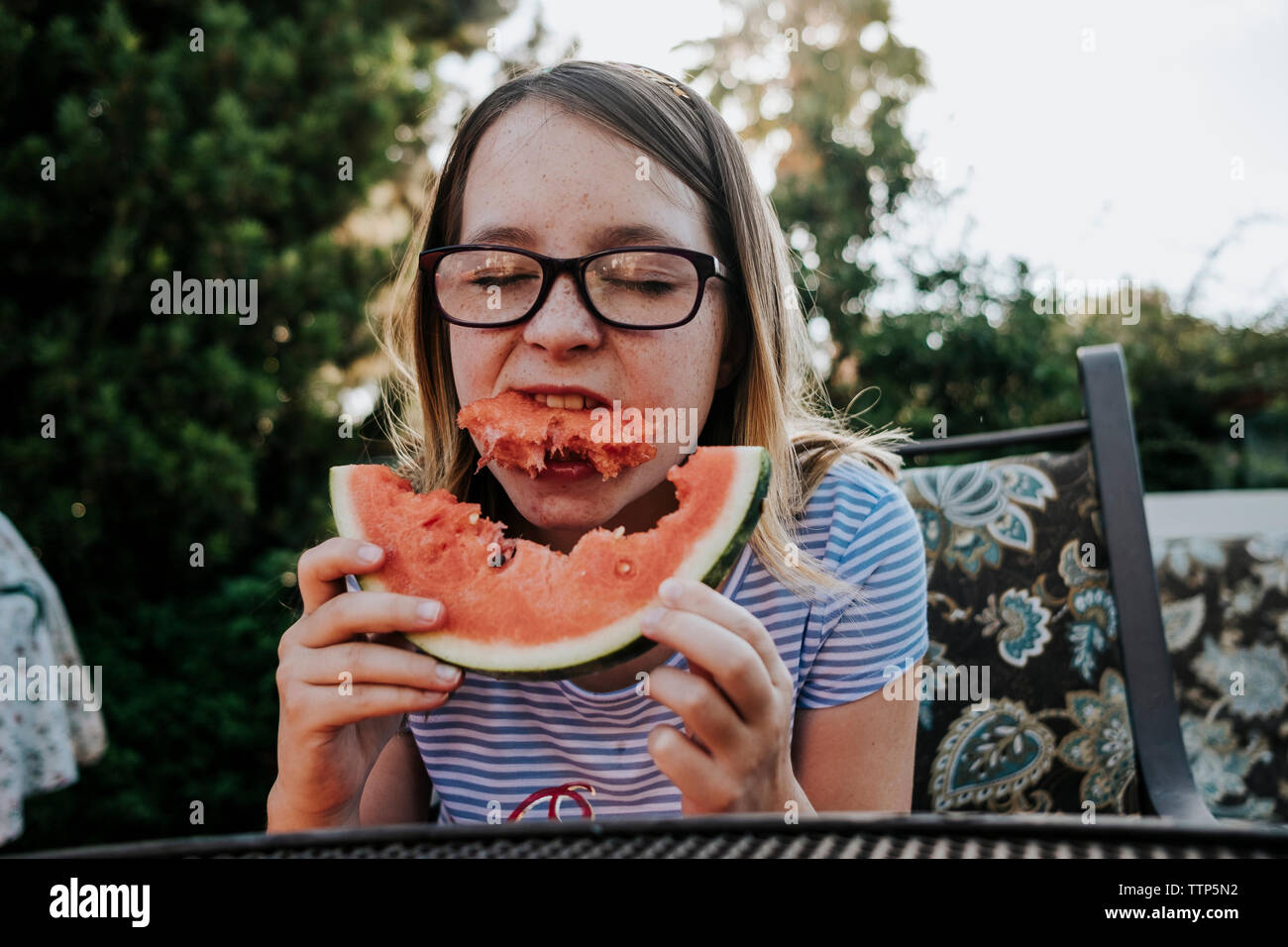 Ragazza indossando occhiali mangiando anguria stando seduti su una sedia contro gli alberi in cantiere Foto Stock