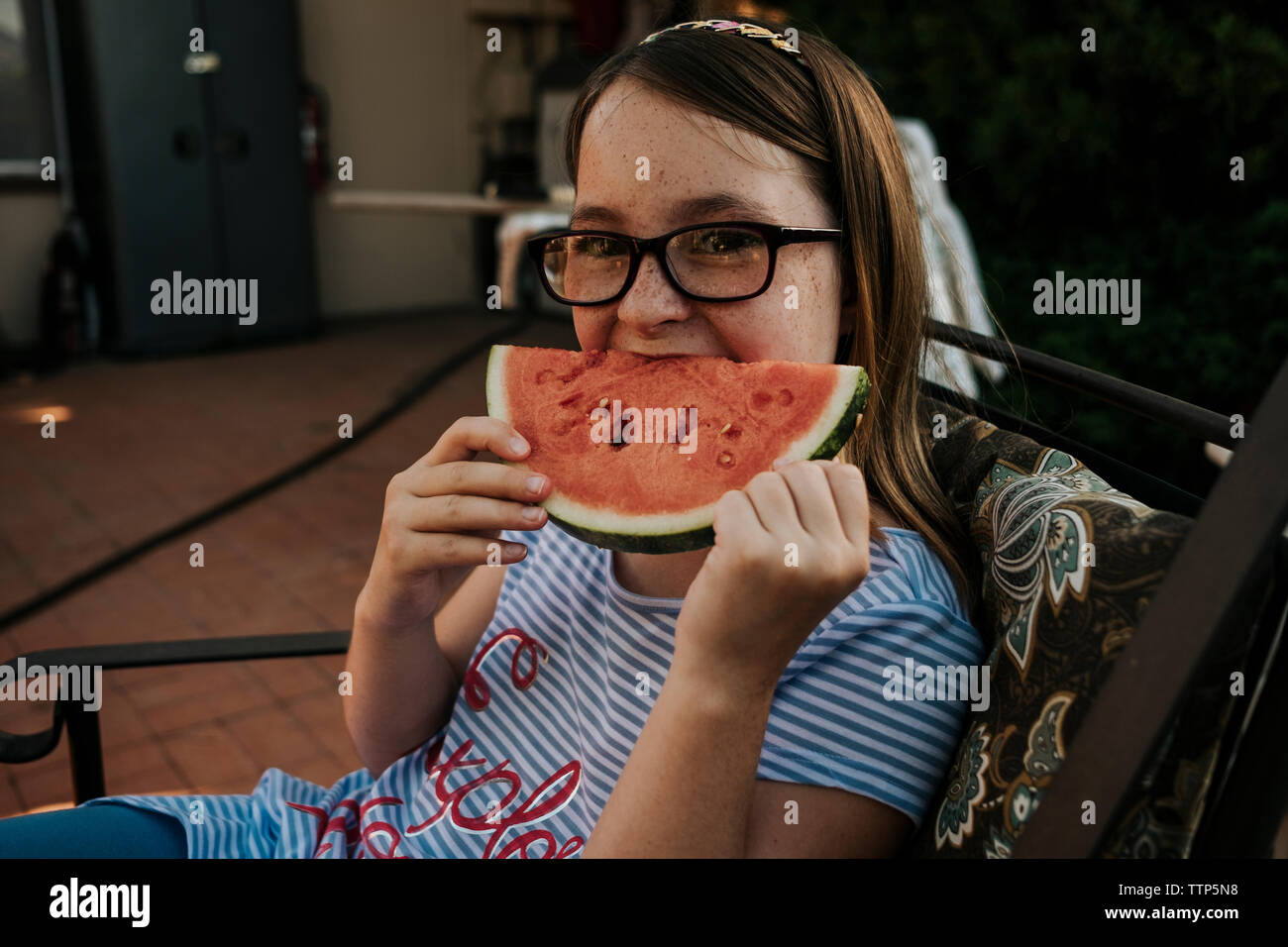 Ritratto di una ragazza che indossa occhiali mangiando anguria mentre è seduto su una sedia in giardino Foto Stock