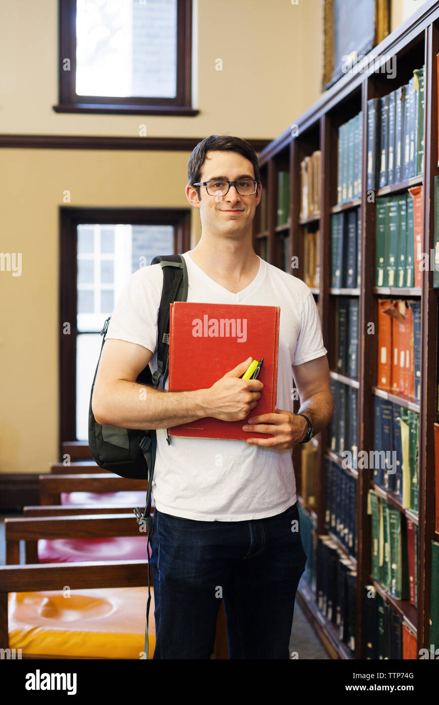 Ritratto di fiducia degli studenti di sesso maschile con zaino azienda prenota permanente, mentre per scaffale in libreria Foto Stock