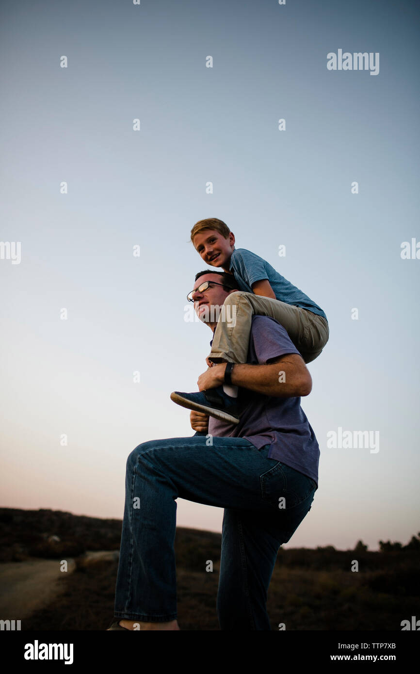 Basso angolo di visione del padre figlio che porta sulle spalle contro il cielo chiaro durante il tramonto Foto Stock
