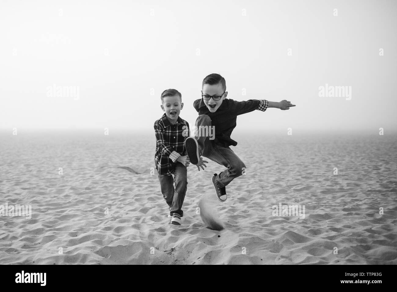 Giocoso fratelli salto sulla sabbia in spiaggia contro il cielo durante la nebbia meteo Foto Stock