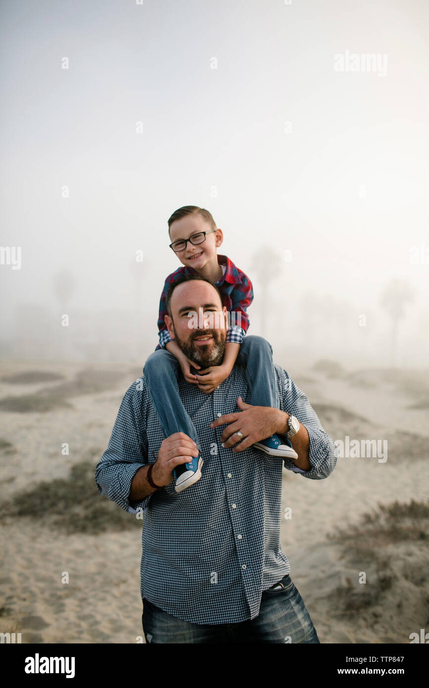 Ritratto di padre che trasportano carino figlio sulle spalle mentre si sta in piedi in spiaggia contro il cielo durante la nebbia meteo Foto Stock