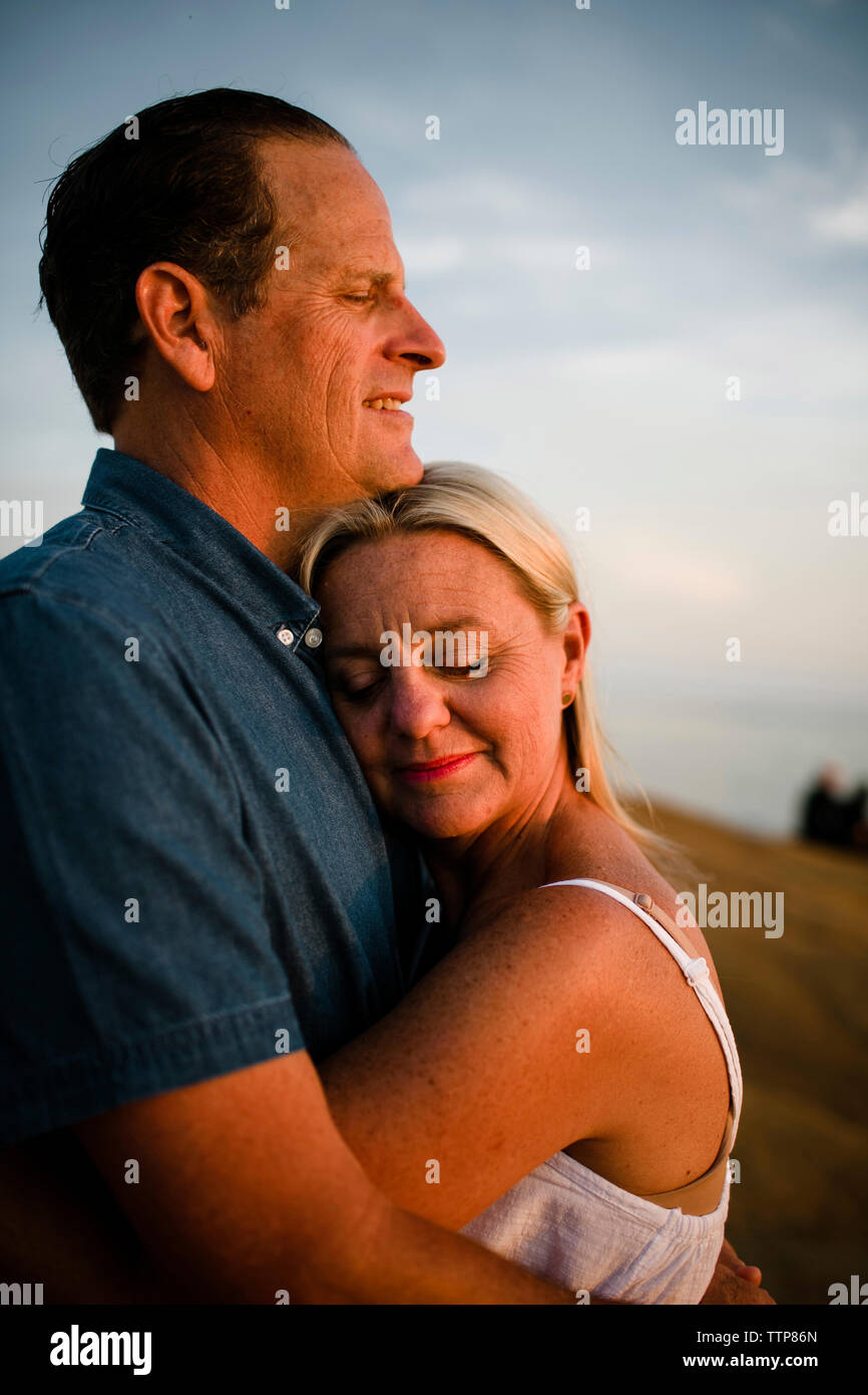Vista laterale della coppia romantica con gli occhi chiusi abbracciando mentre si sta in piedi sul dirupo contro il cielo durante il tramonto Foto Stock
