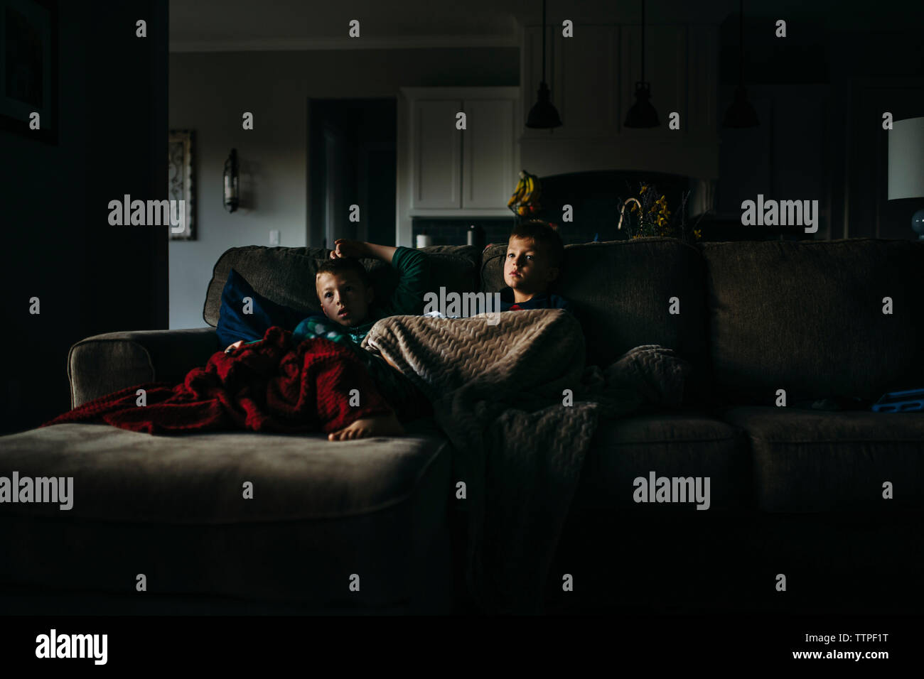 Fratelli sotto le coperte a guardare la TV seduti sul divano di casa in camera oscura Foto Stock