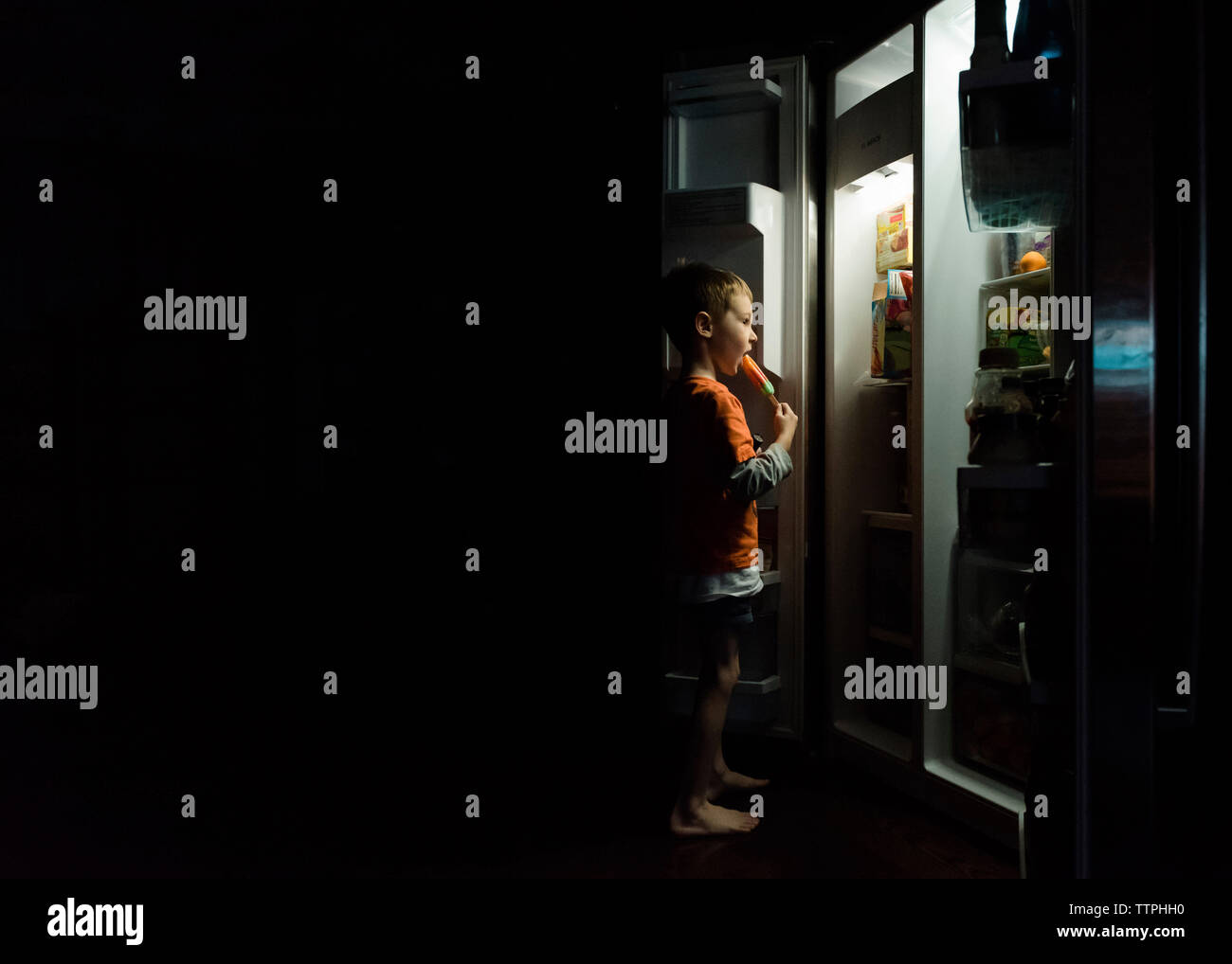 Boy eating ice pop mentre in piedi nella parte anteriore del frigorifero a casa Foto Stock