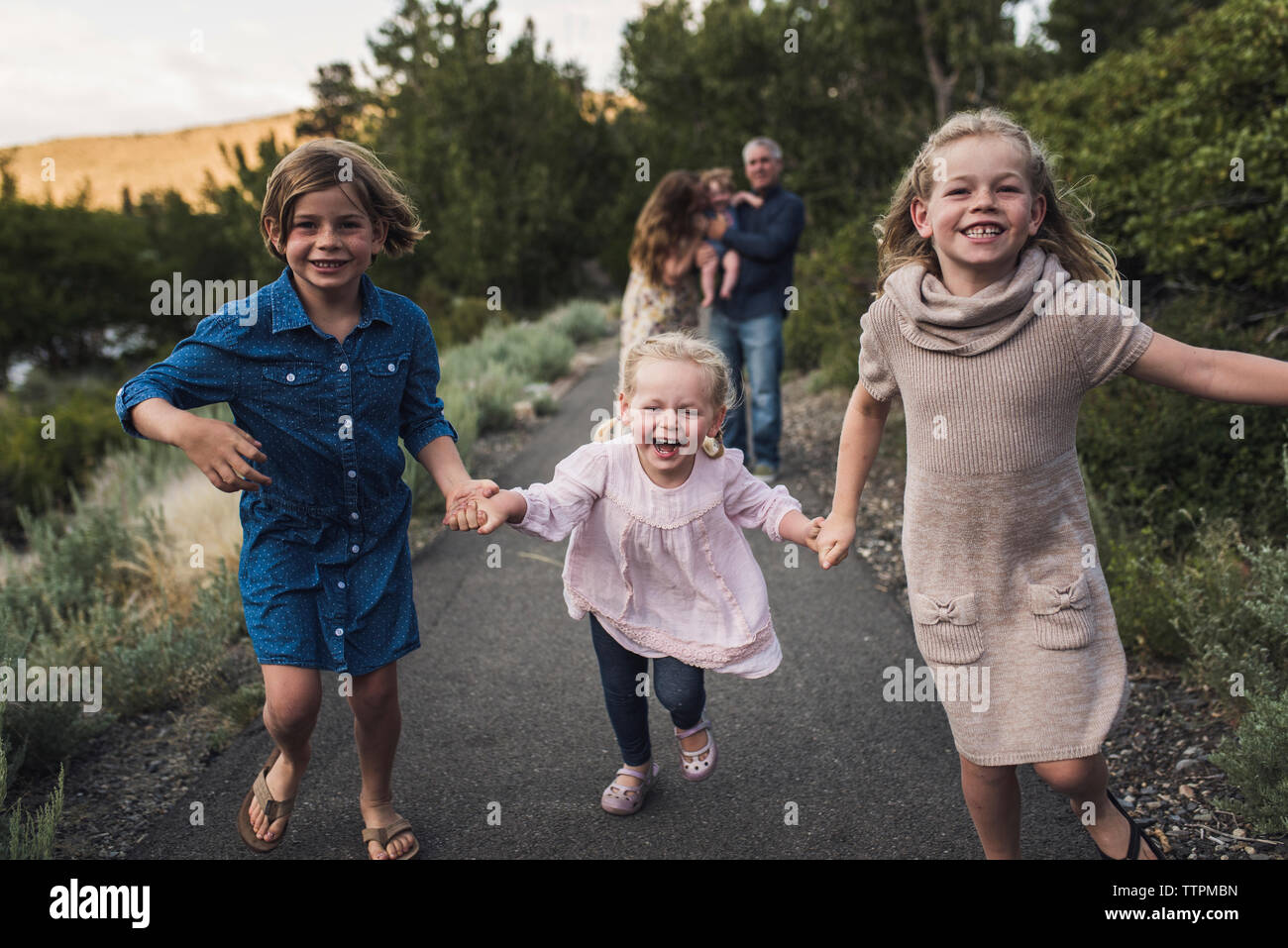 Felice sorelle tenendo le mani durante il funzionamento su strada contro i genitori nella foresta Foto Stock