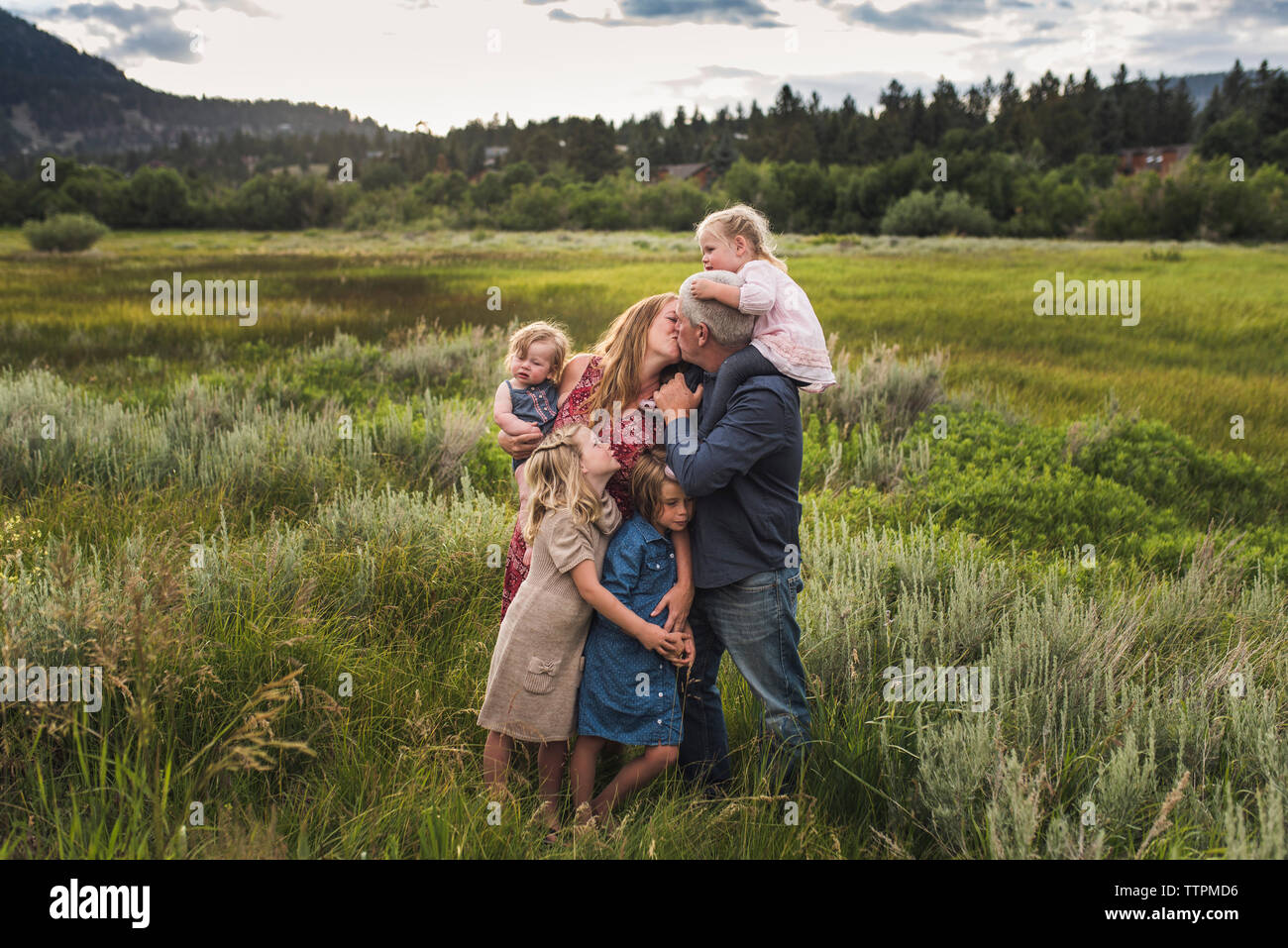 I genitori romantica baciare stando in piedi con le figlie sul campo erboso contro sky in foresta Foto Stock