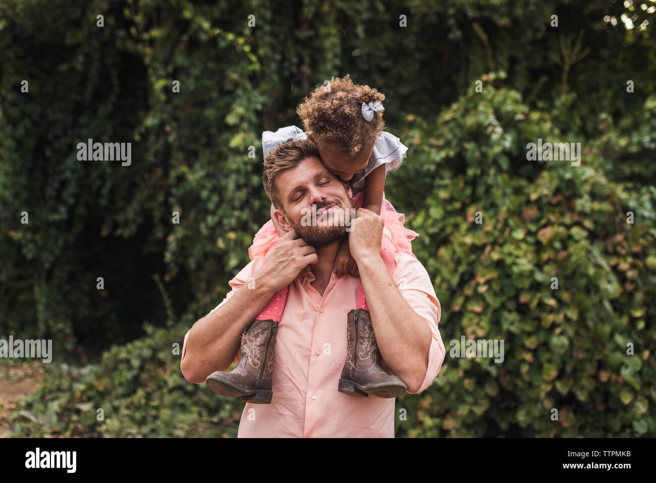Padre con gli occhi chiusi che trasportano figlia sulle spalle mentre in piedi contro piante in posizione di parcheggio Foto Stock