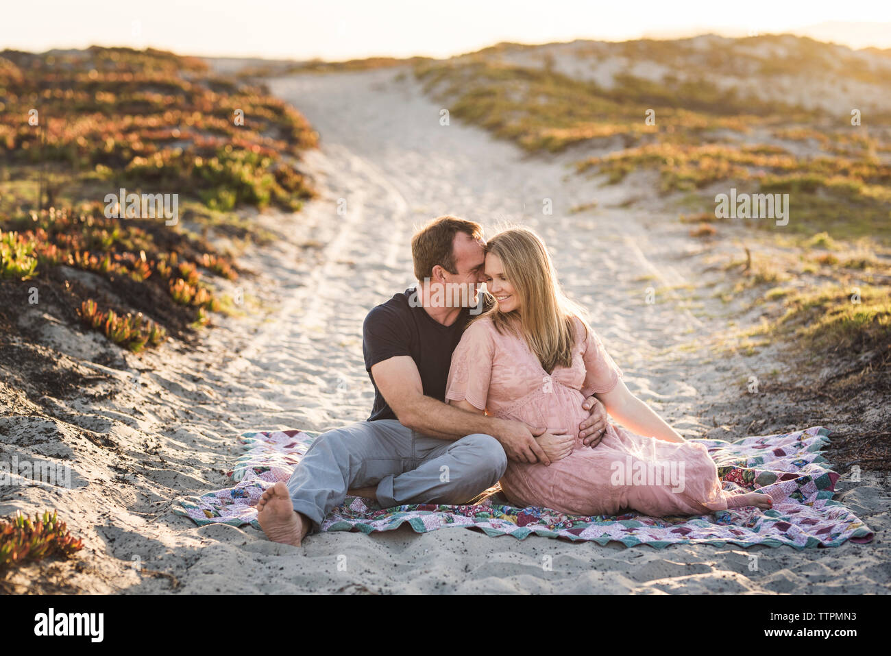 Romantica Coppia sorridente seduto su una coperta a beach contro il cielo chiaro durante il tramonto Foto Stock