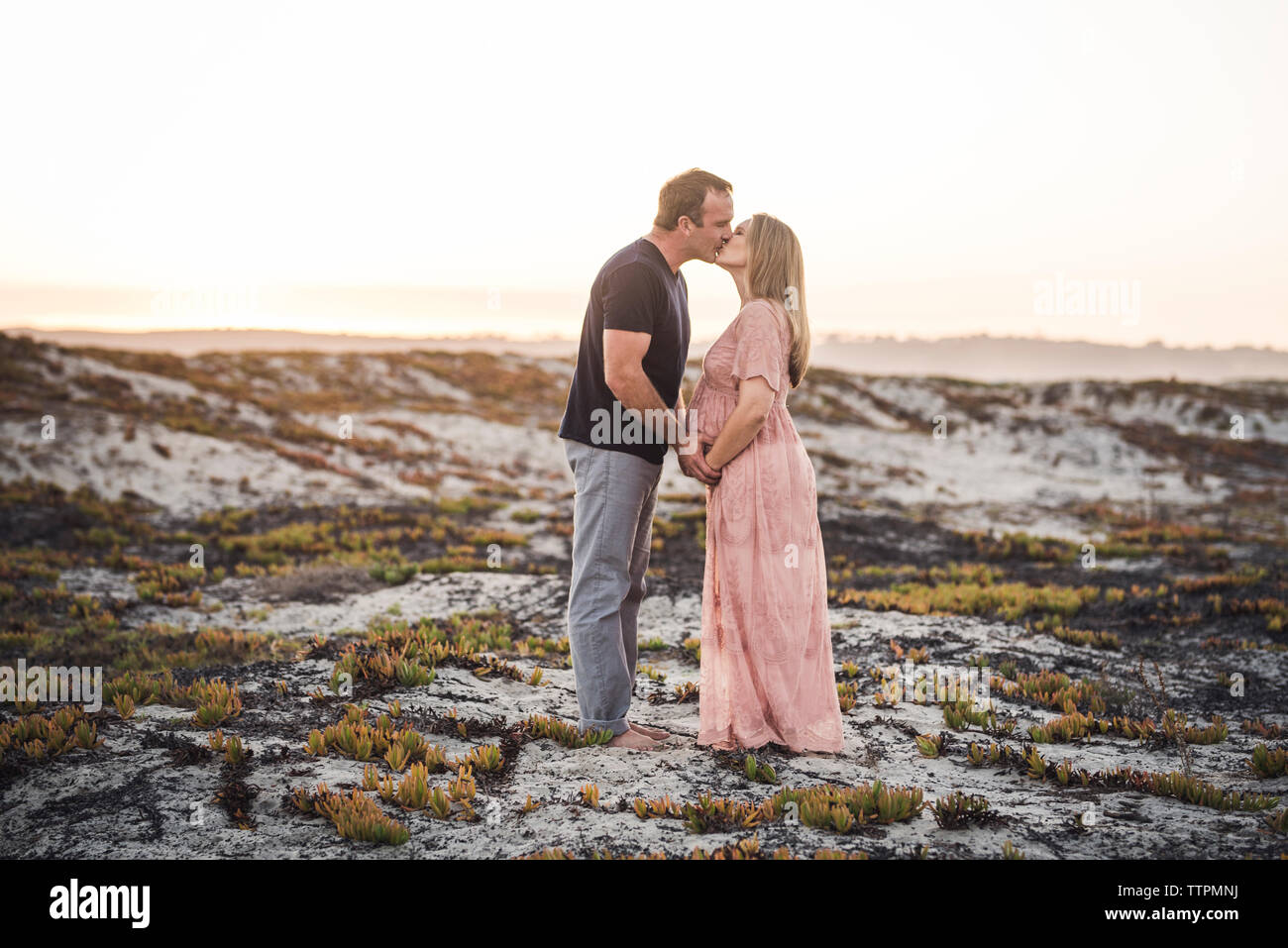 Vista laterale della coppia romantica baciare mentre permanente sulla spiaggia contro il cielo chiaro durante il tramonto Foto Stock
