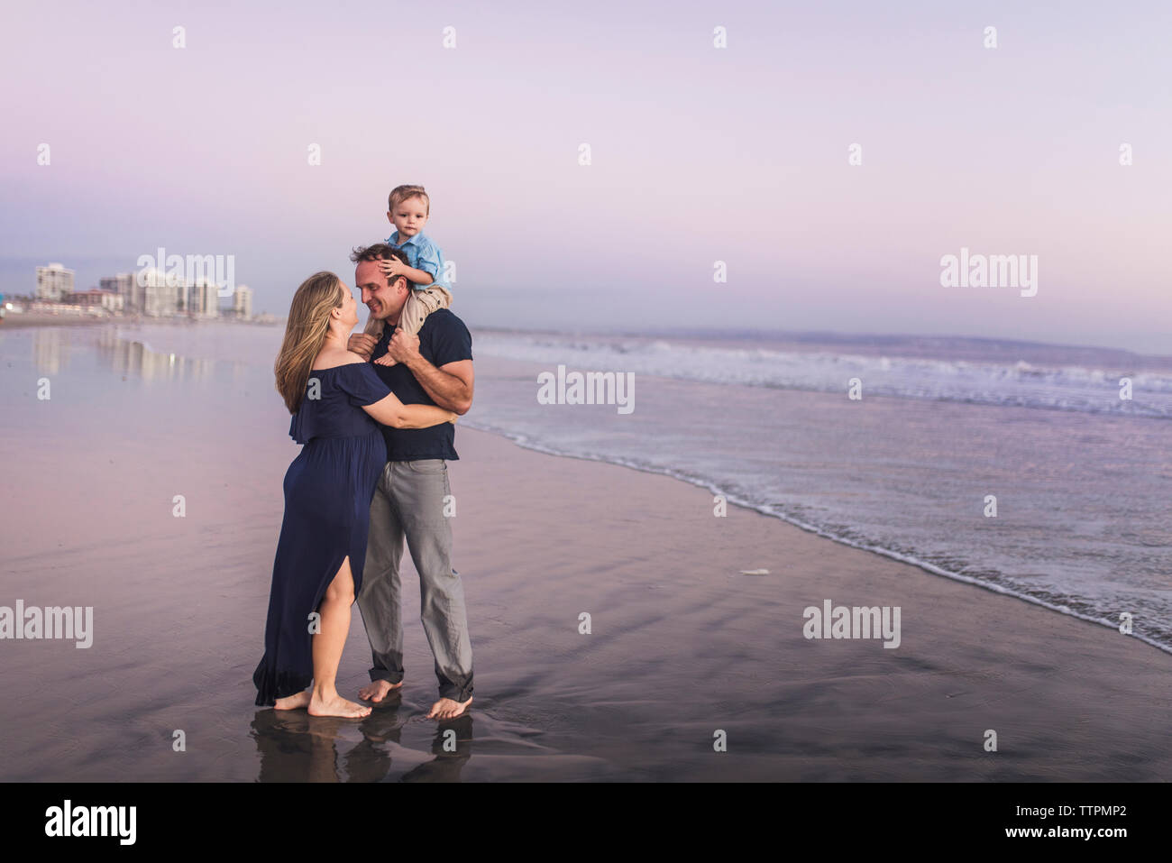 Romantico marito guardando mia moglie mentre trasportano figlio sulla spiaggia contro il cielo durante il tramonto Foto Stock