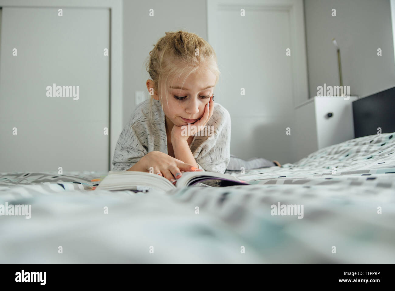 La ragazza con la mano sul mento libro lettura mentre giaceva sul letto di casa Foto Stock