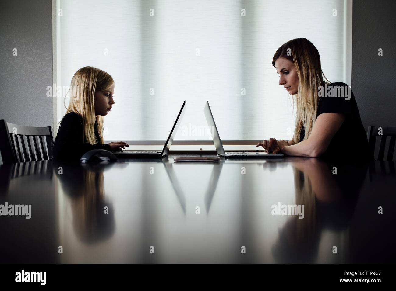 Vista laterale della madre e figlia utilizzando computer portatili sul tavolo mentre è seduto a casa Foto Stock