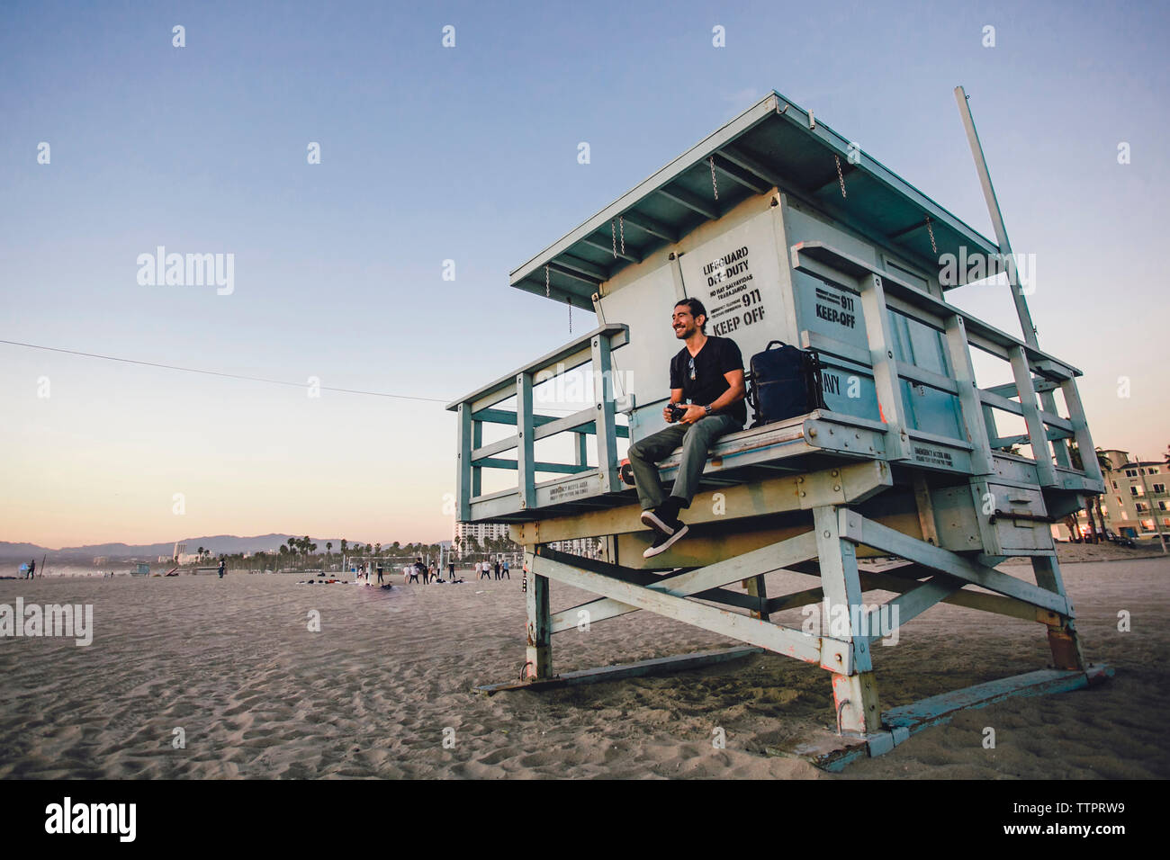 Lunghezza completa di uomo seduto su lifeguard hut in spiaggia durante il tramonto Foto Stock