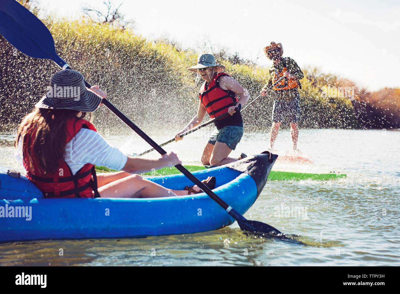 Gli amici gli spruzzi di acqua sulla donna seduta in kayak gonfiabili mentre paddleboarding al lago Foto Stock