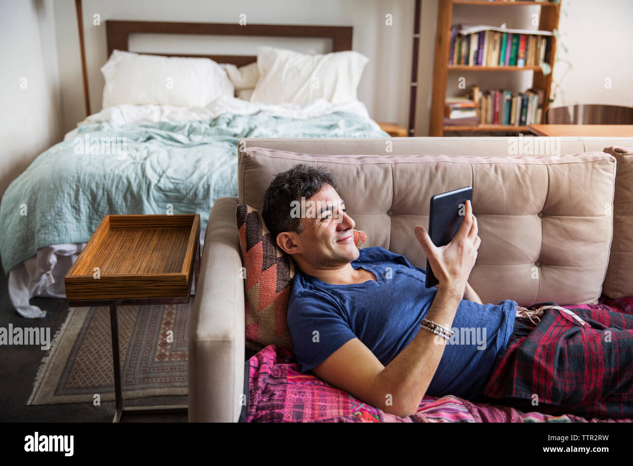 Uomo sorridente utilizzando computer tavoletta mentre sdraiato sul divano di casa Foto Stock