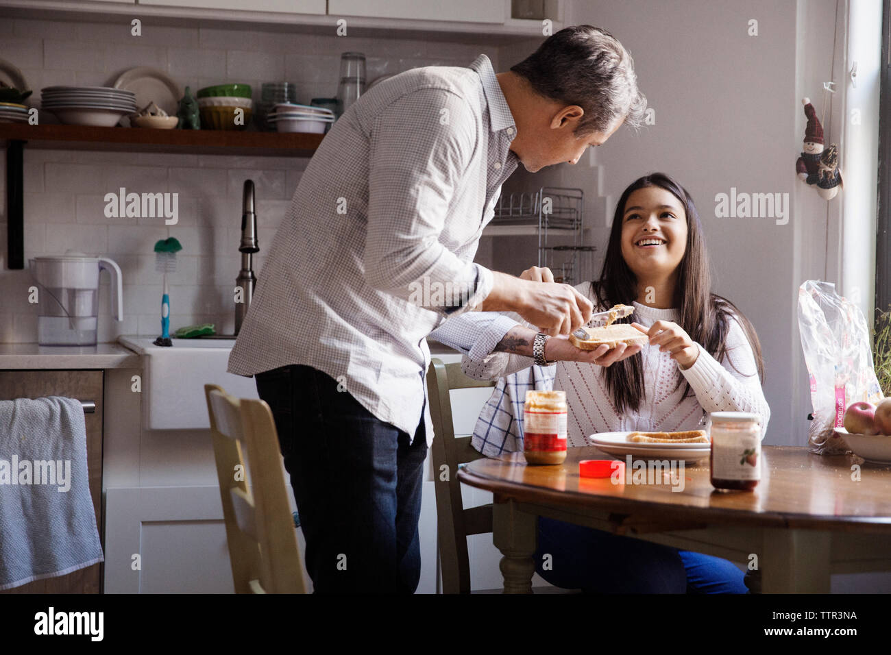 Ragazza sorridente guardando padre applicare burro di arachidi sul pane in cucina Foto Stock