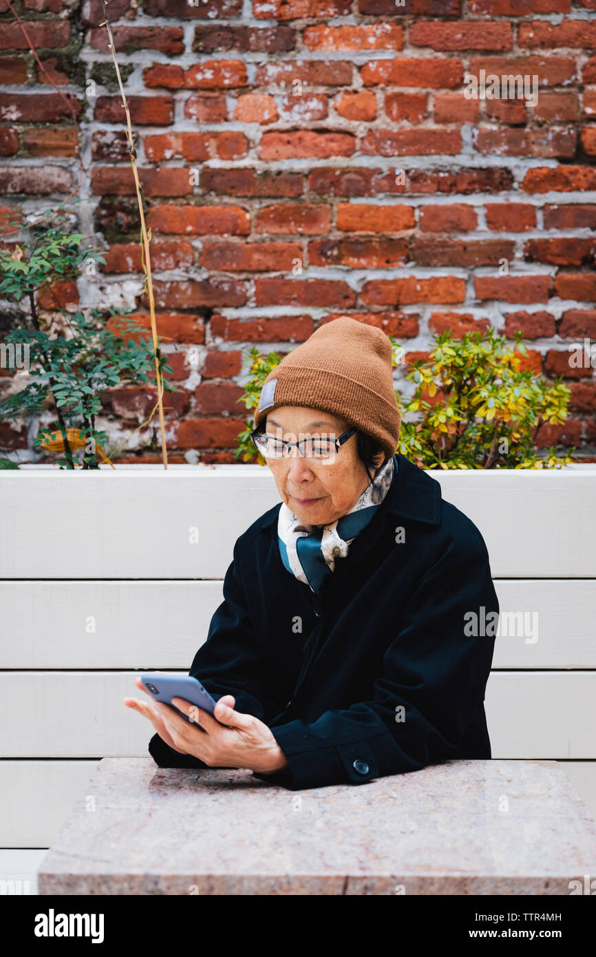 Senior donna che indossa abiti caldi utilizzando smart phone mentre è seduto sulla panchina contro il muro di mattoni in città Foto Stock