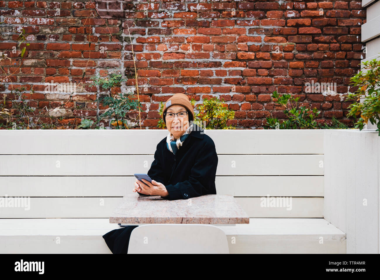 Ritratto di donna senior indossando un abbigliamento caldo utilizzando smart phone mentre è seduto sulla panchina contro il muro di mattoni in città Foto Stock