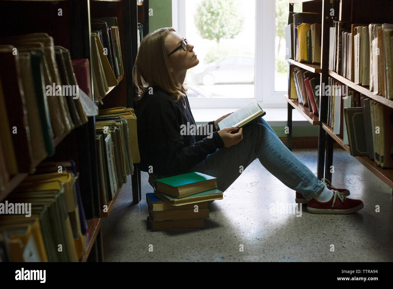 Donna che mantiene libro e guardando verso l'alto mentre è seduto nella libreria Foto Stock