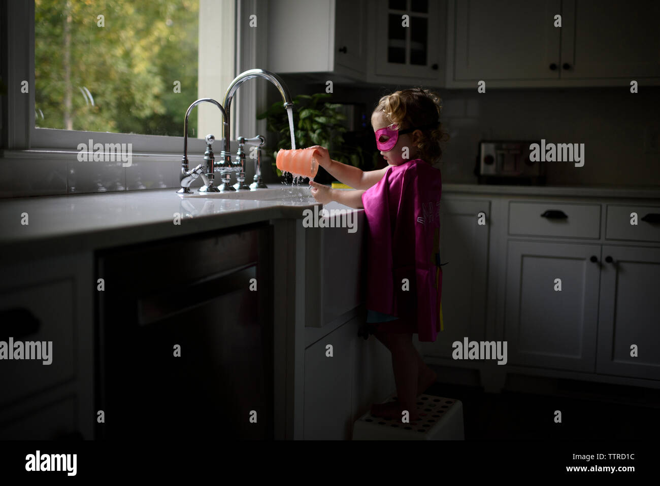 Vista laterale della ragazza di indossare il costume del supereroe di lavarsi le mani nel lavello da cucina mentre in piedi a casa Foto Stock