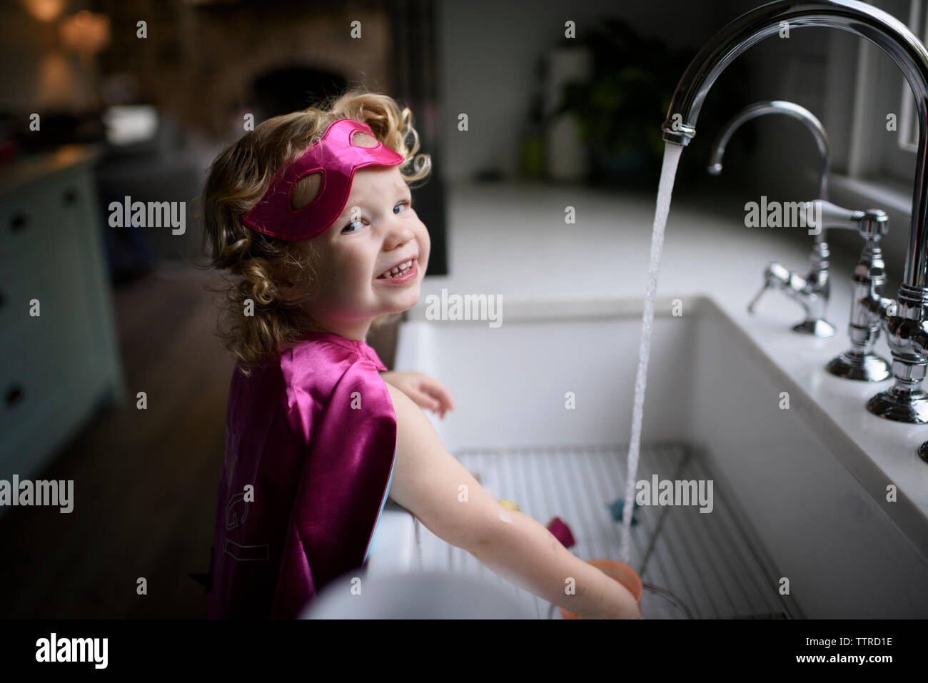 Ritratto di ragazza sorridente indossando il costume del supereroe di lavarsi le mani nel lavello da cucina mentre in piedi a casa Foto Stock