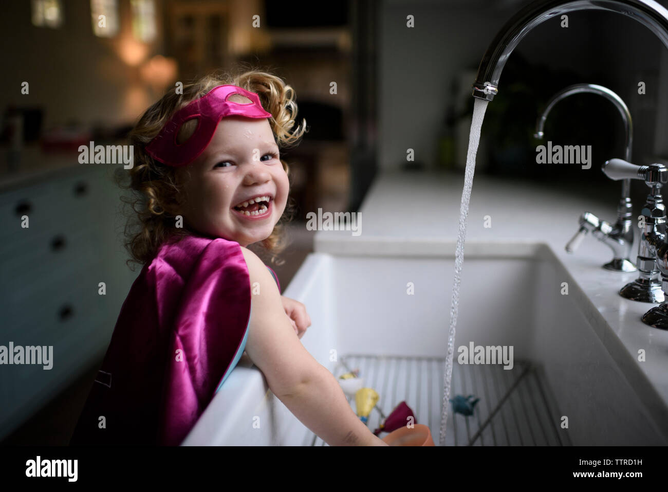 Ritratto di ragazza sorridente indossando il costume del supereroe di lavarsi le mani nel lavello da cucina a casa Foto Stock