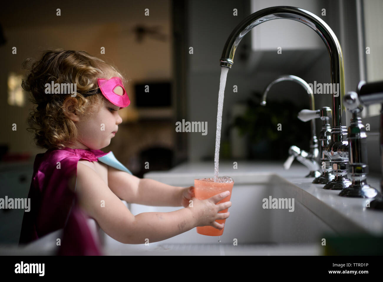 Vista laterale della ragazza di indossare il costume di supereroi il riempimento di acqua nel bicchiere mentre in piedi a casa Foto Stock