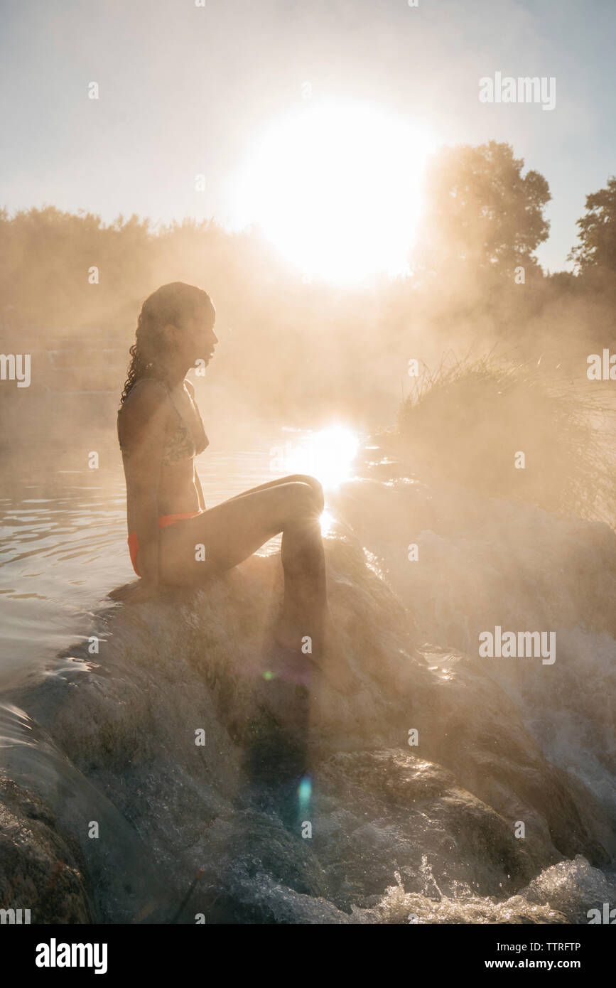 Vista laterale della donna seduta sulle rocce in emissione di vapore Piscina termale durante la giornata di sole Foto Stock