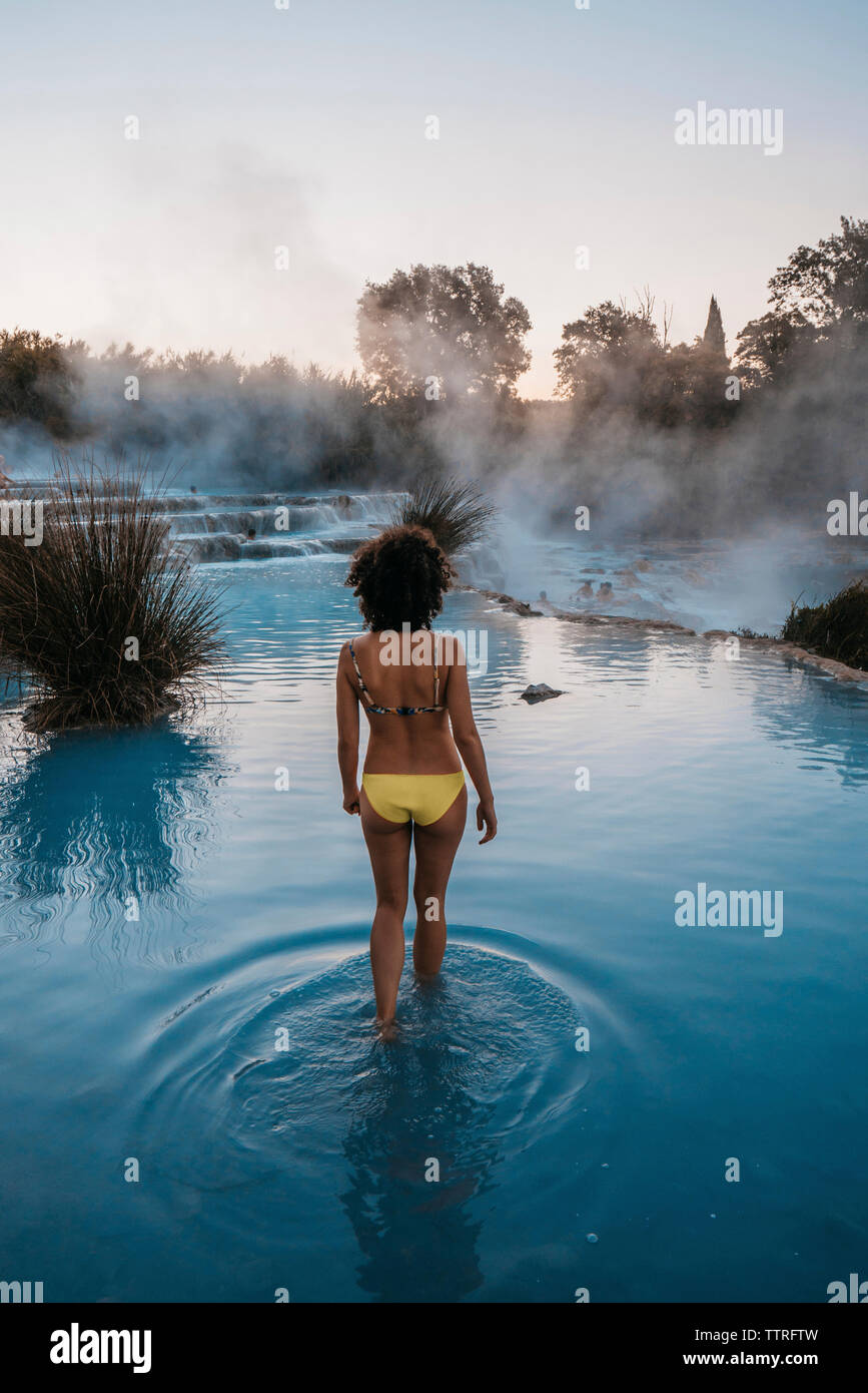 Vista posteriore della donna che indossa un bikini mentre si cammina in piscina termale durante il tramonto Foto Stock