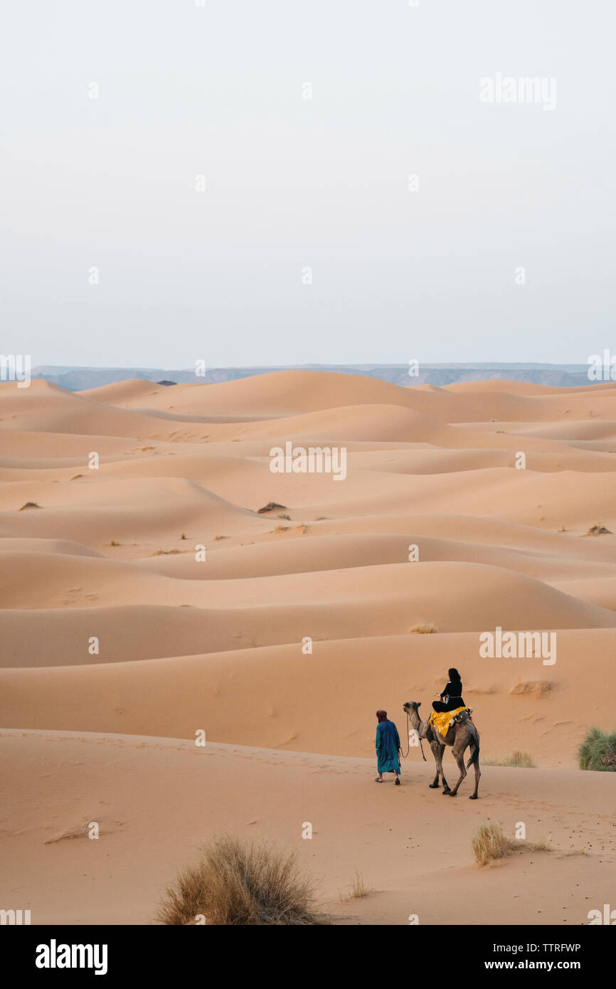 Vista posteriore di una donna in sella a dorso di cammello al deserto del Sahara contro il cielo chiaro Foto Stock