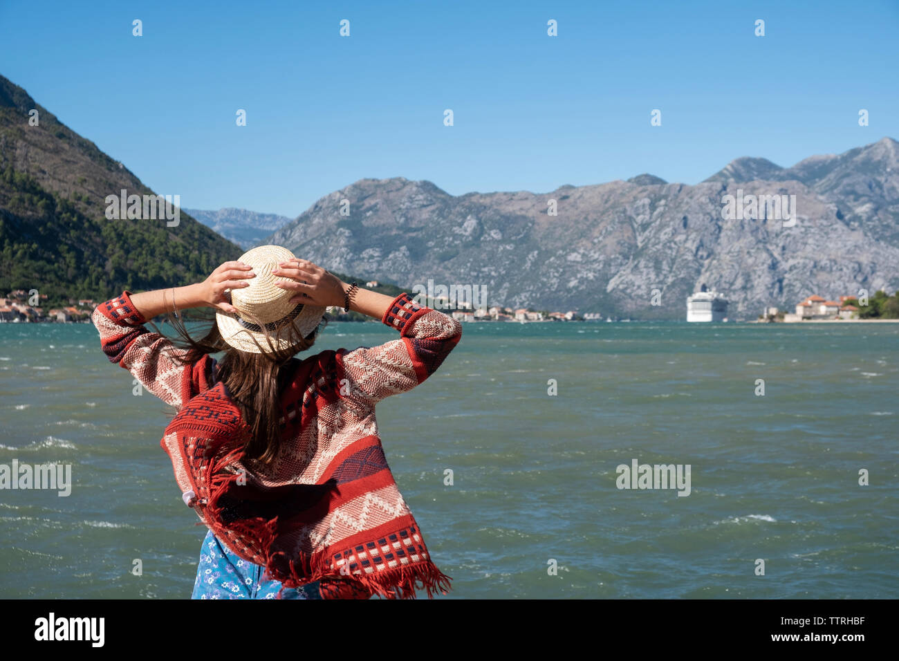 Vista posteriore della donna che indossa il cappello con braccia alzate guardando il mare mentre in piedi contro il cielo blu e chiaro durante la giornata di sole Foto Stock