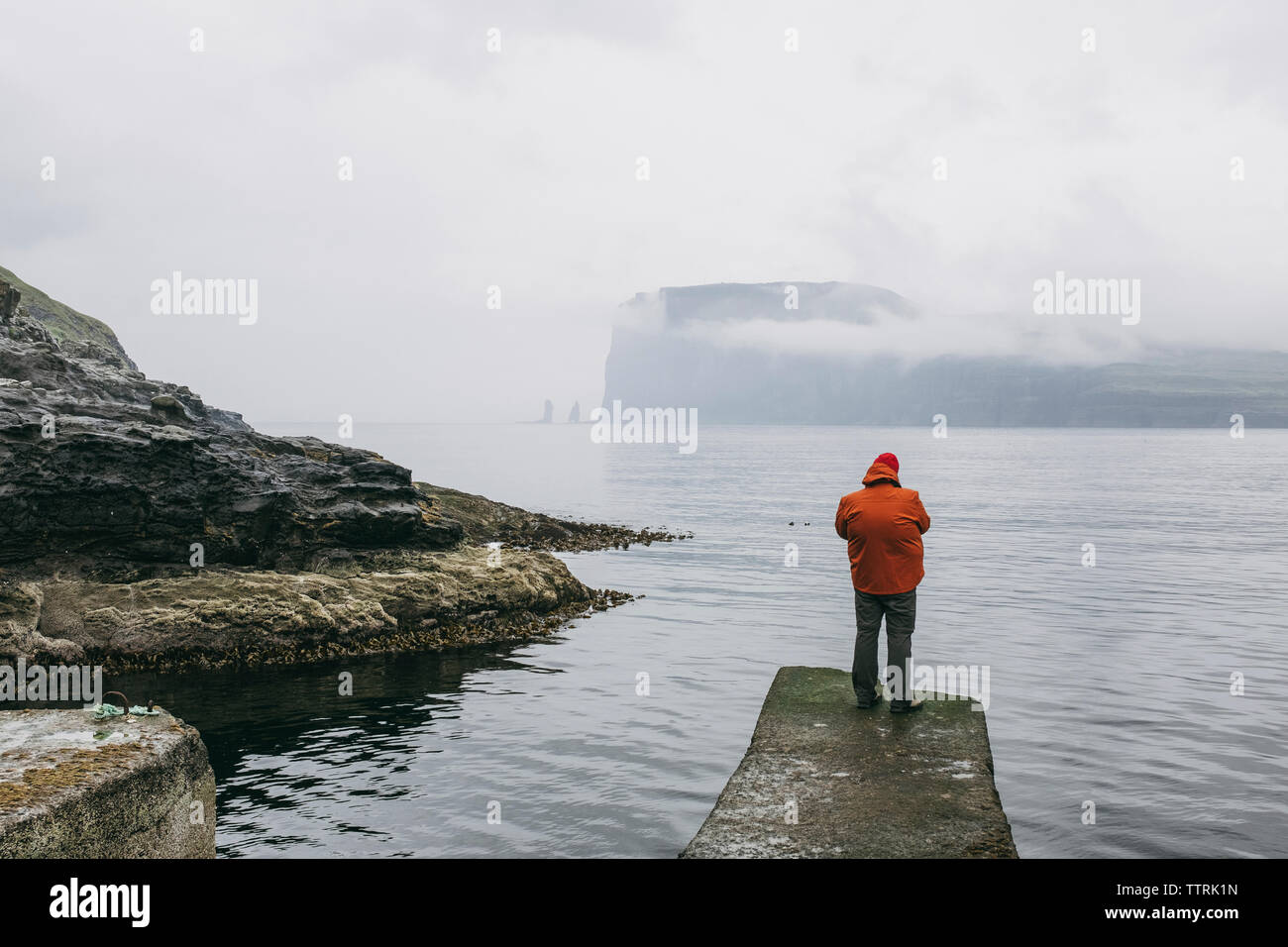 Vista posteriore di un escursionista in giacca con cappuccio in piedi sul molo contro il mare durante la nebbia meteo Foto Stock