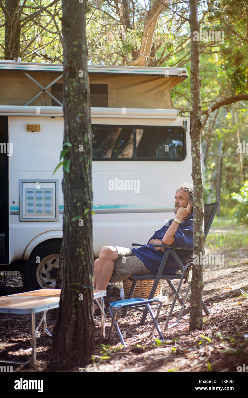 Riflessivo senior uomo seduto sulla sedia pieghevole contro il rimorchio di viaggio nella foresta Foto Stock