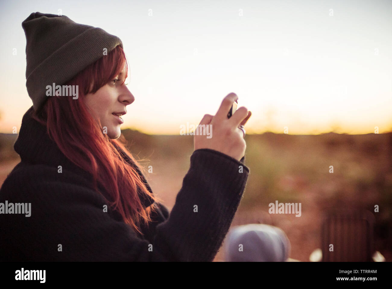 Vista laterale della donna fotografando attraverso smart phone contro sky durante il tramonto Foto Stock