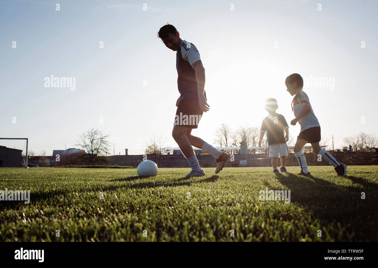 Basso angolo di visione dell uomo che giocano a calcio con i bambini sul campo erboso contro il cielo chiaro durante il tramonto Foto Stock