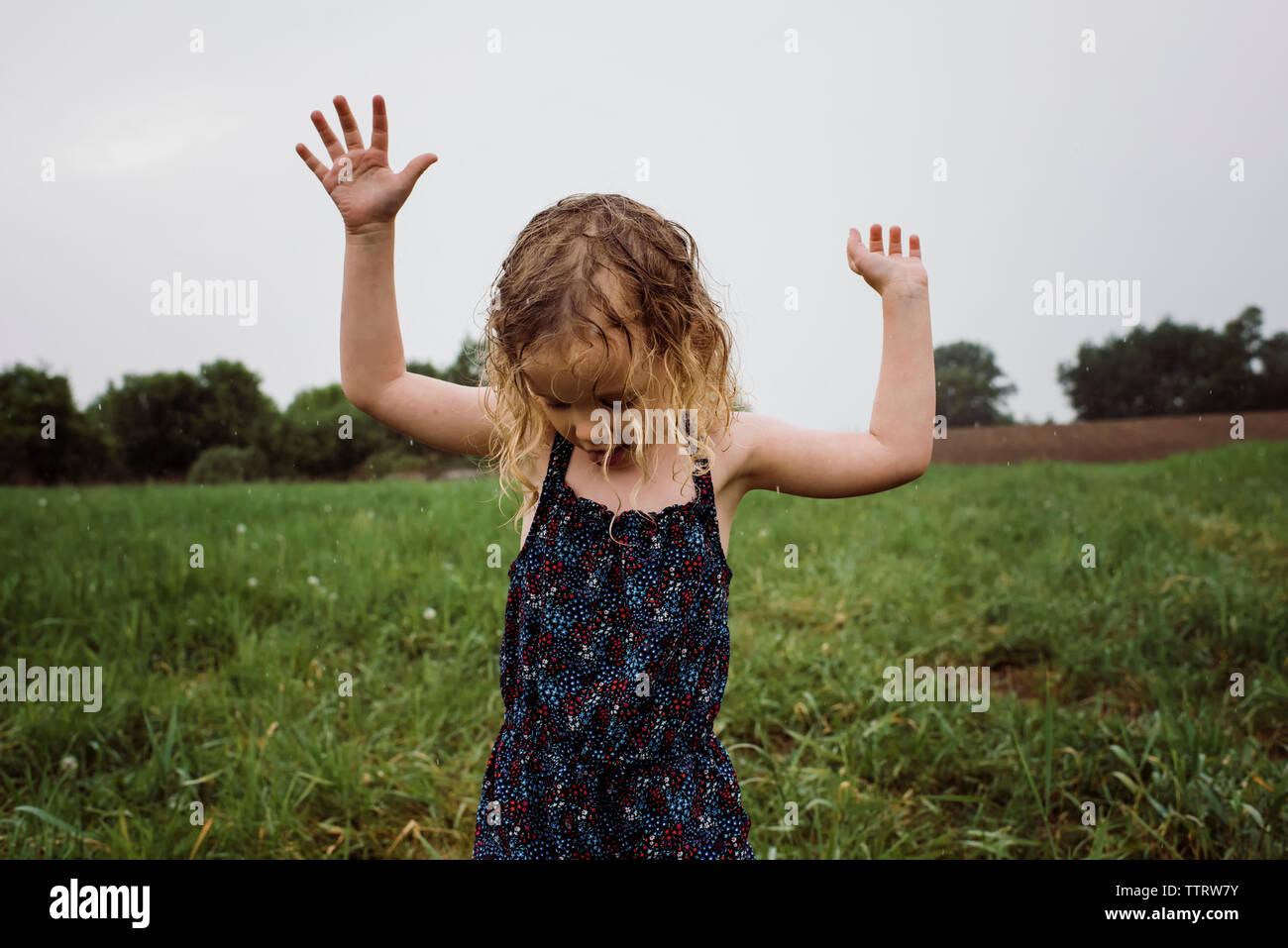 Wet ragazza con braccia alzate in piedi sul campo erboso contro il cielo al parco durante la stagione delle piogge Foto Stock