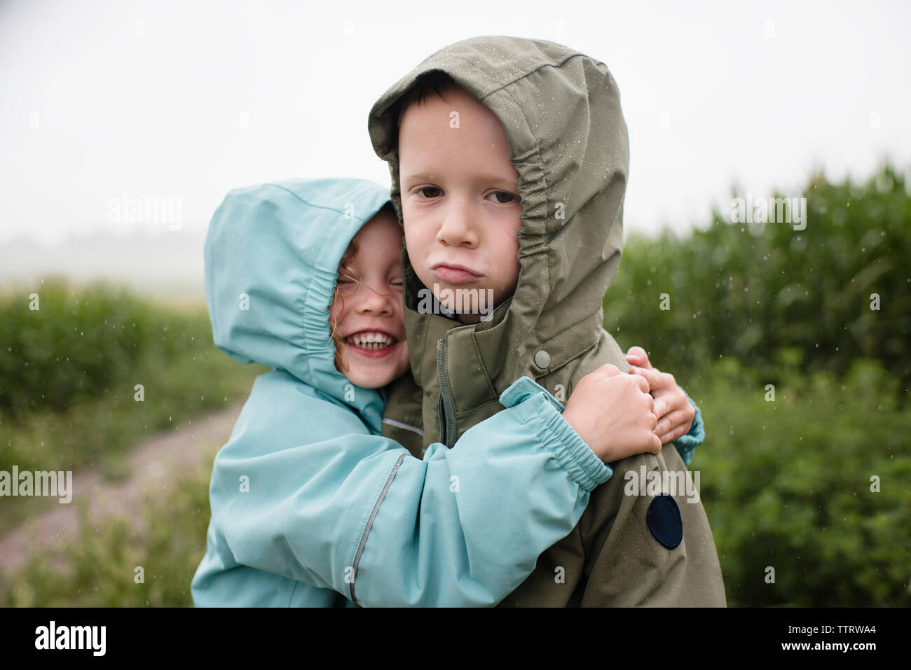 Ritratto di triste fratello essendo abbracciato da felice sorella mentre in piedi contro piante durante la stagione delle piogge Foto Stock