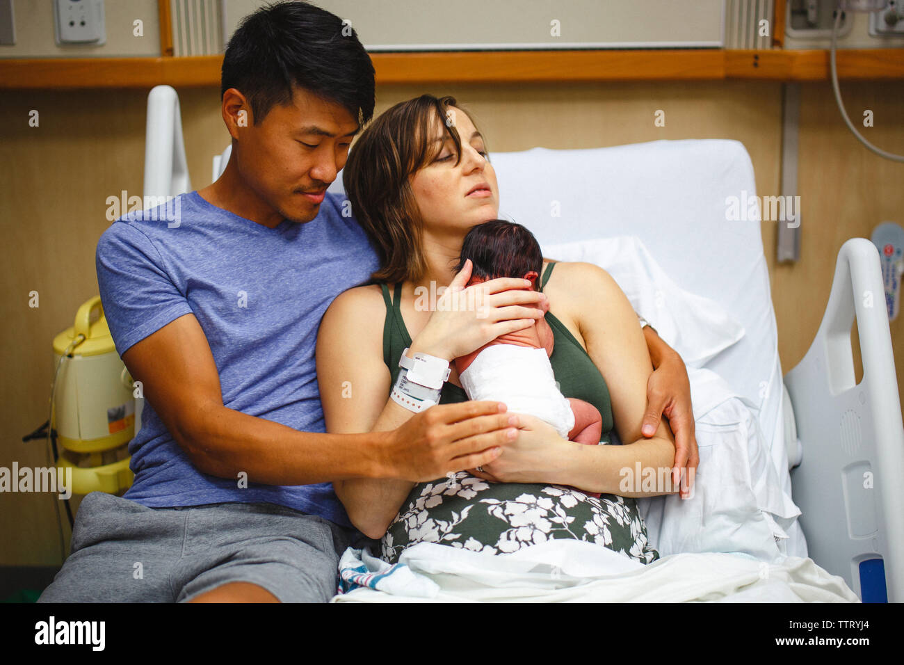 Una nuova mamma riposa in un letto di ospedale cullano il neonato, marito a sid Foto Stock
