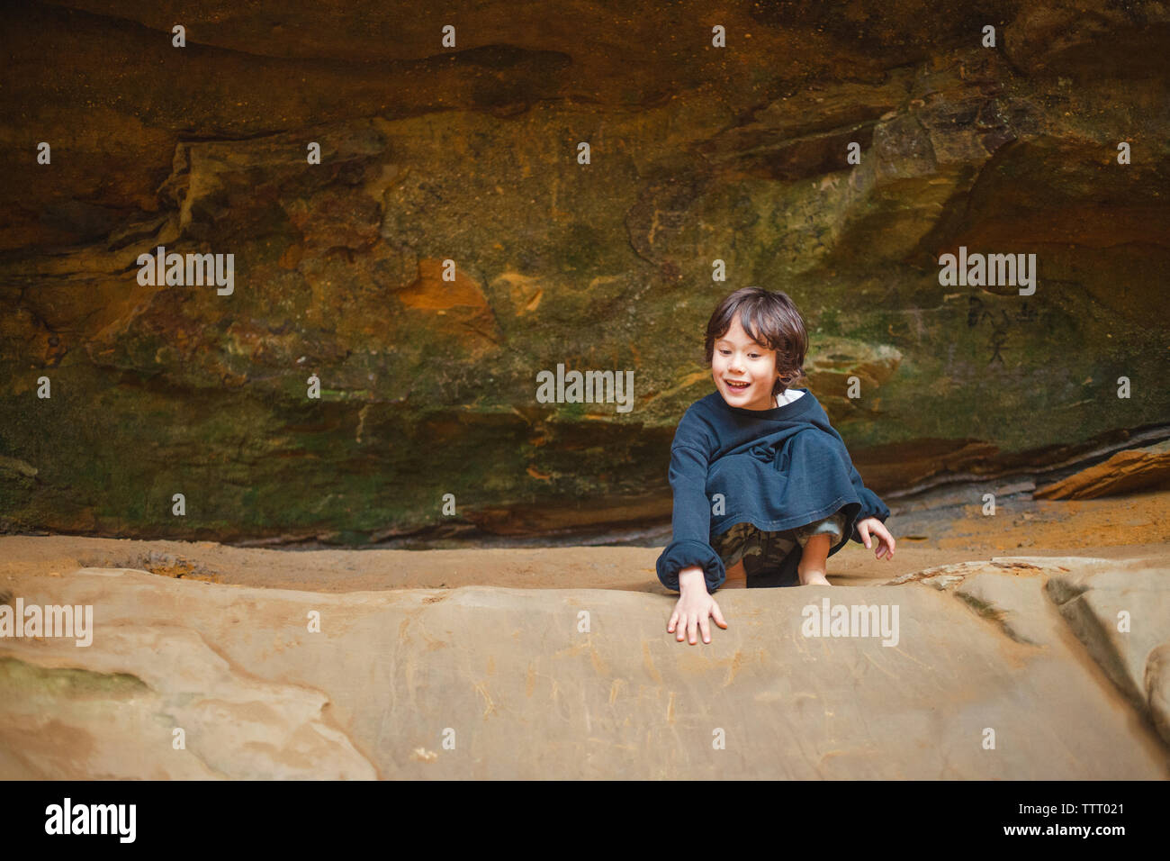 Un piccolo bambino felice si accovaccia su una cengia rocciosa contro la scogliera di arenaria Foto Stock