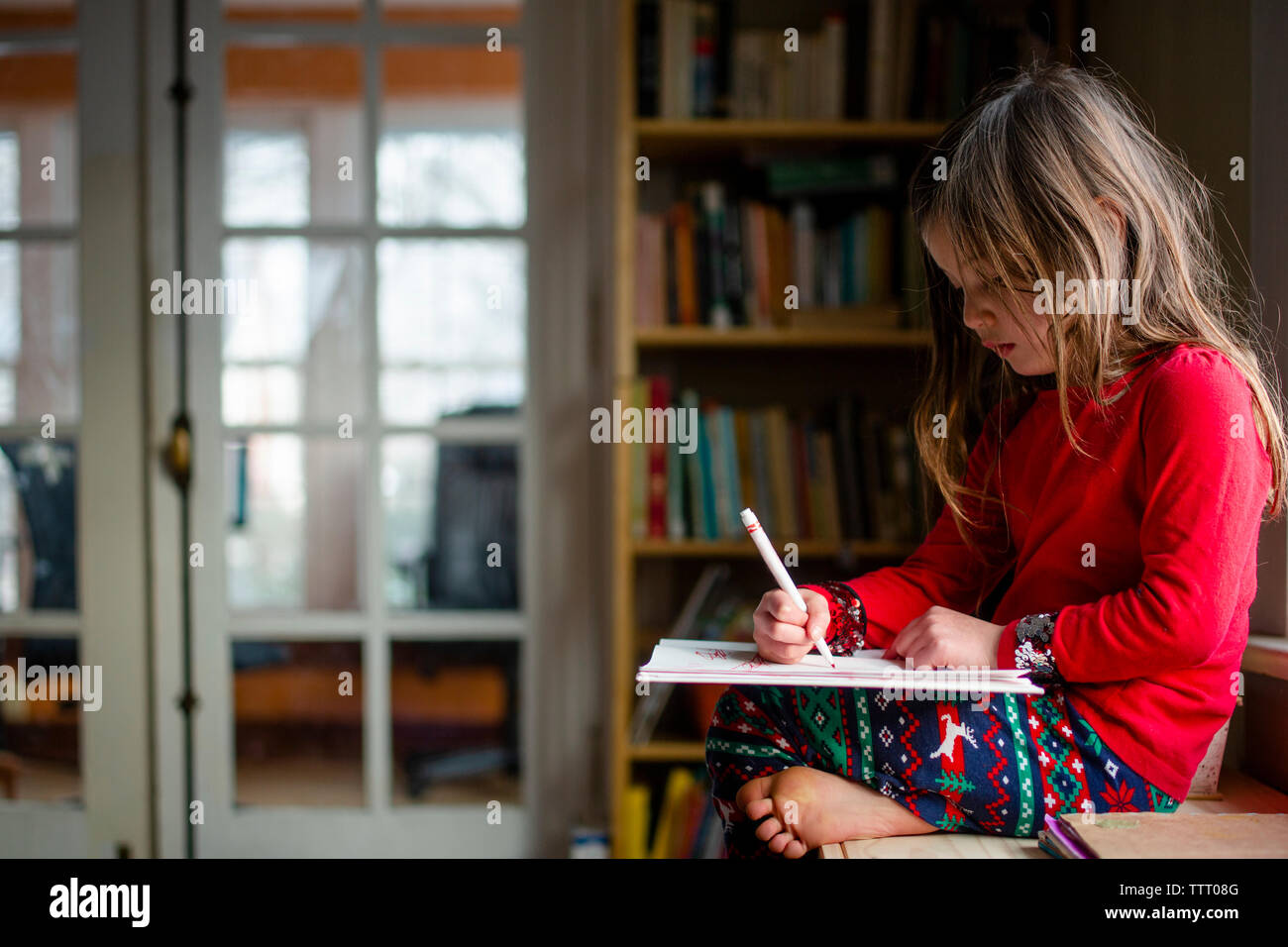 Una piccola ragazza seduta a piedi nudi da una libreria scritta in un notebook Foto Stock