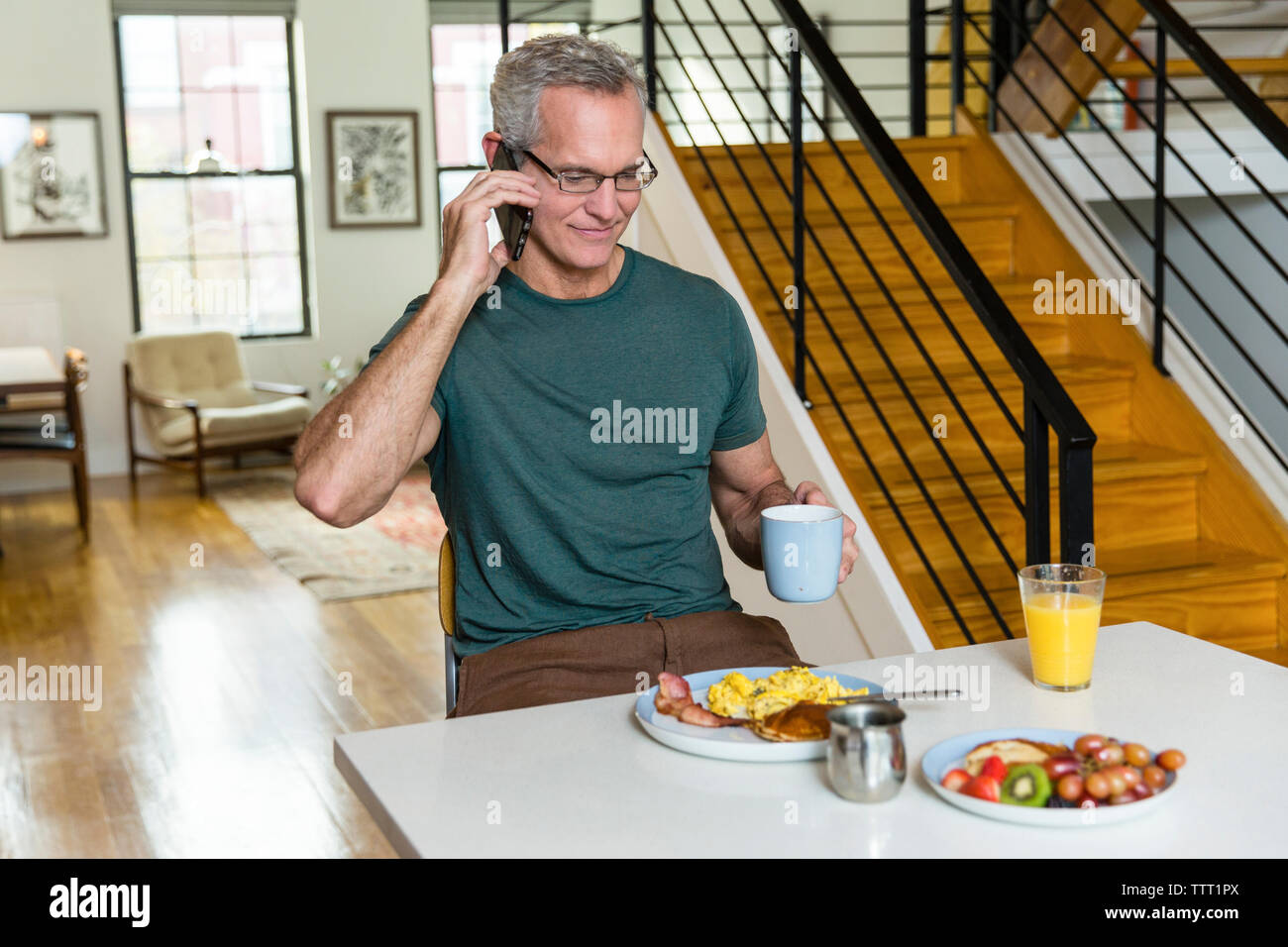 Uomo maturo holding tazza di caffè mentre sta rispondendo alle smart phone al tavolo da pranzo Foto Stock
