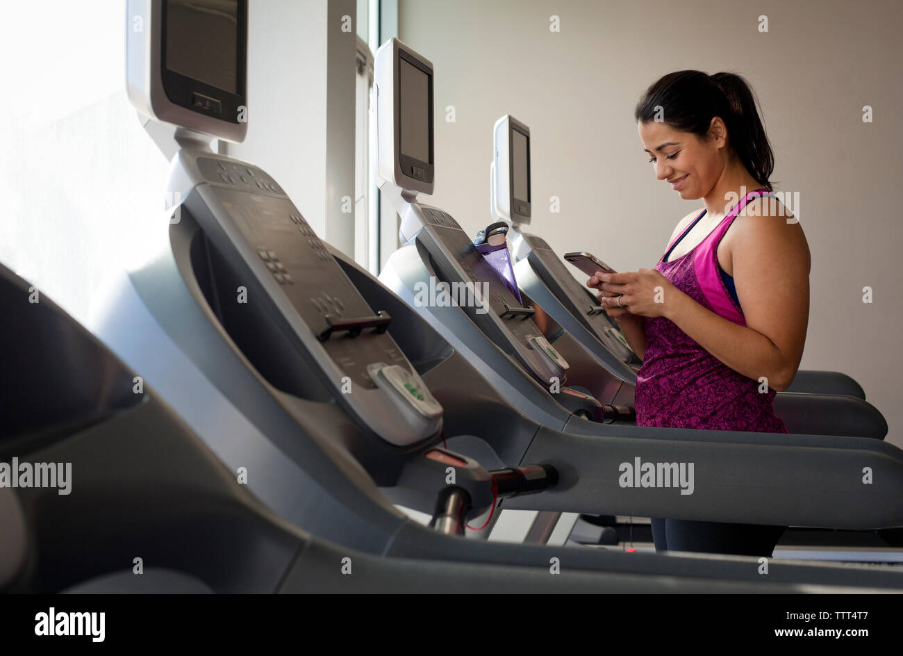 Donna sorridente con smart phone mentre si sta in piedi sul tapis roulant in palestra Foto Stock