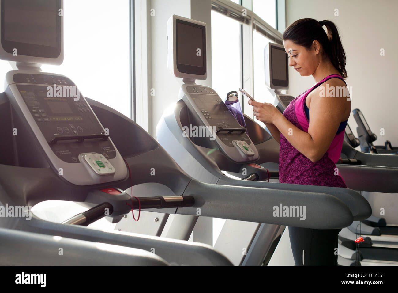 Vista laterale della donna utilizzando smart phone mentre si sta in piedi sul tapis roulant in palestra Foto Stock