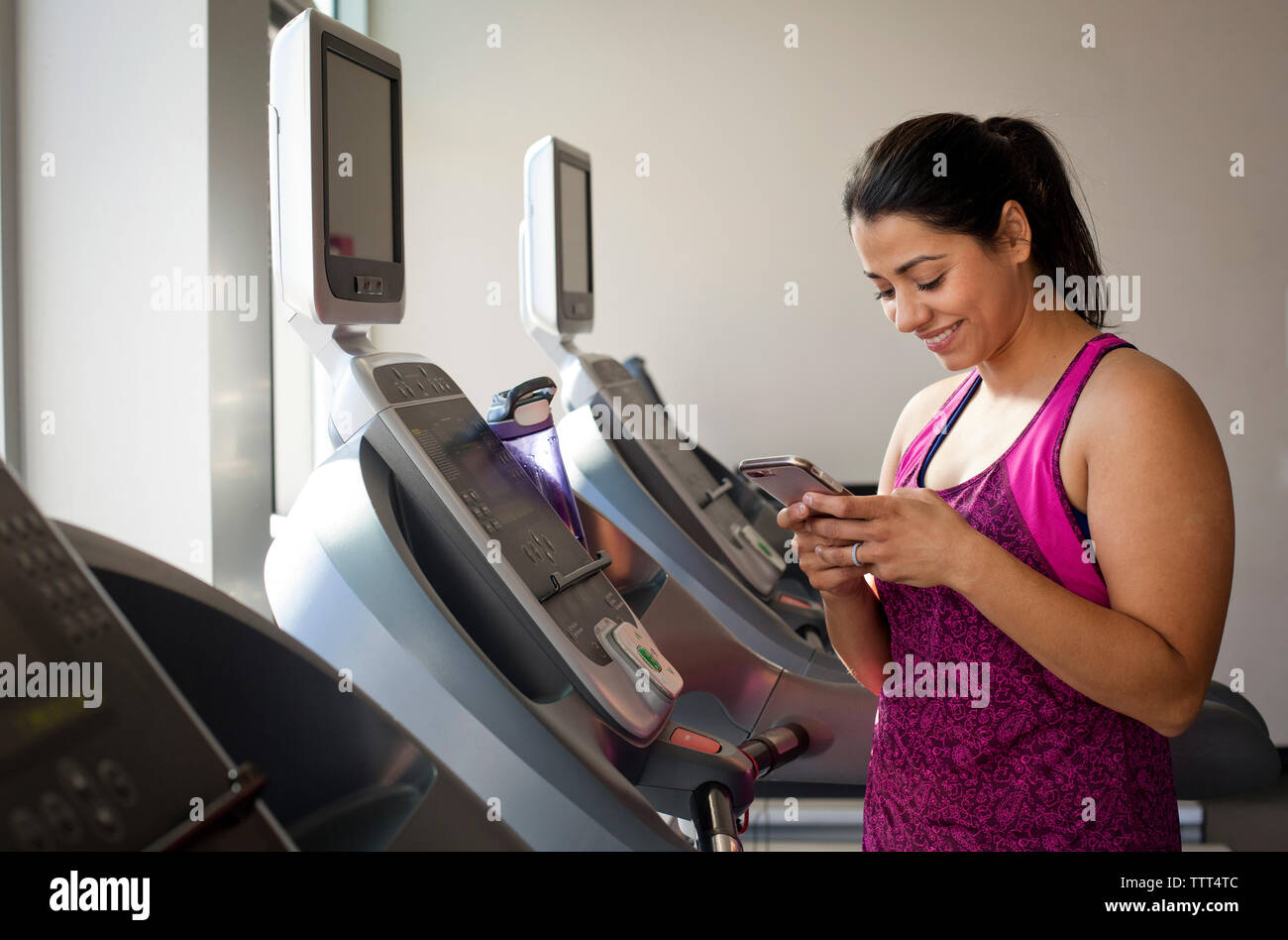 Donna sorridente con smart phone mentre si sta in piedi sul tapis roulant in palestra Foto Stock